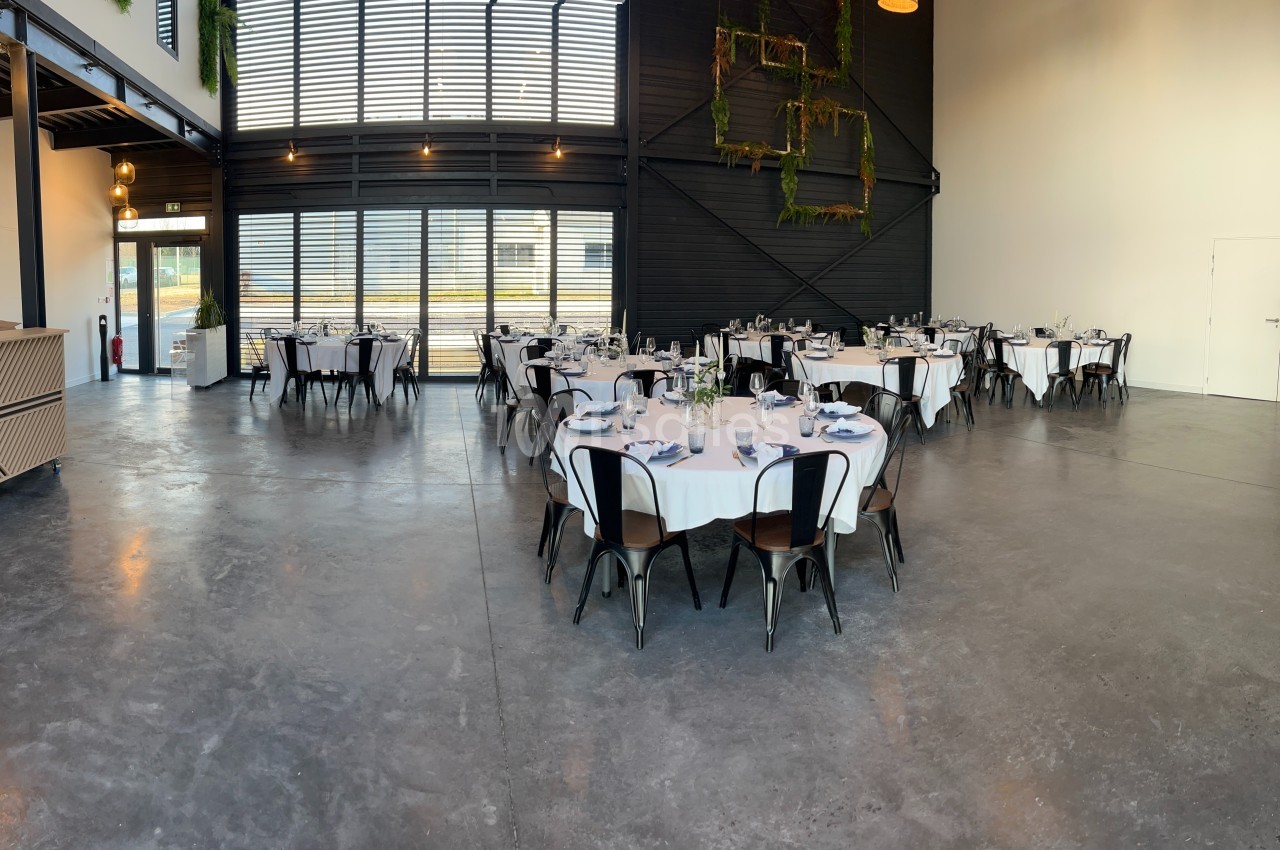 Salle de réception lumineuse avec tables rondes dressées, chaises noires et décorations végétales sur les murs.