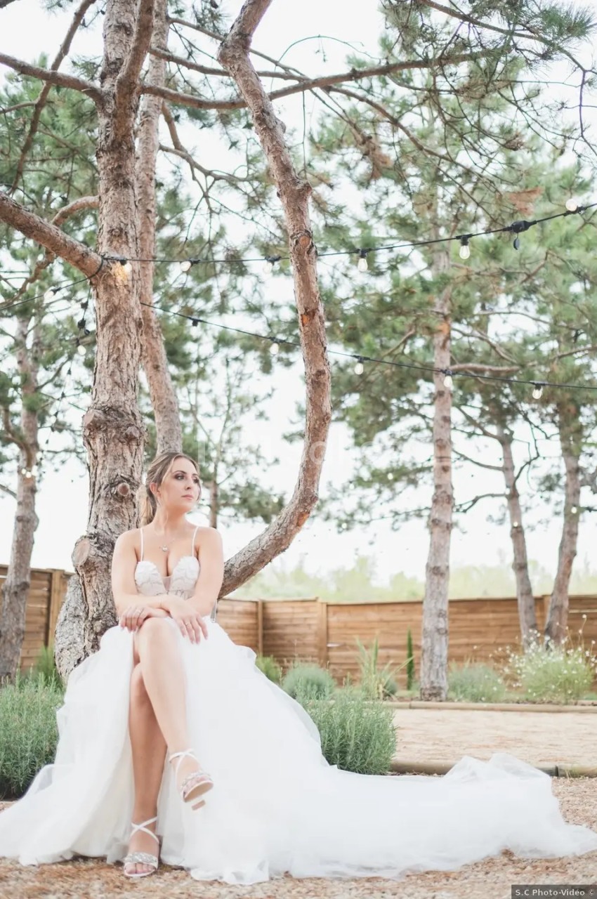 Une femme en robe de mariée blanche est assise près d'un arbre dans un jardin entouré de pins.