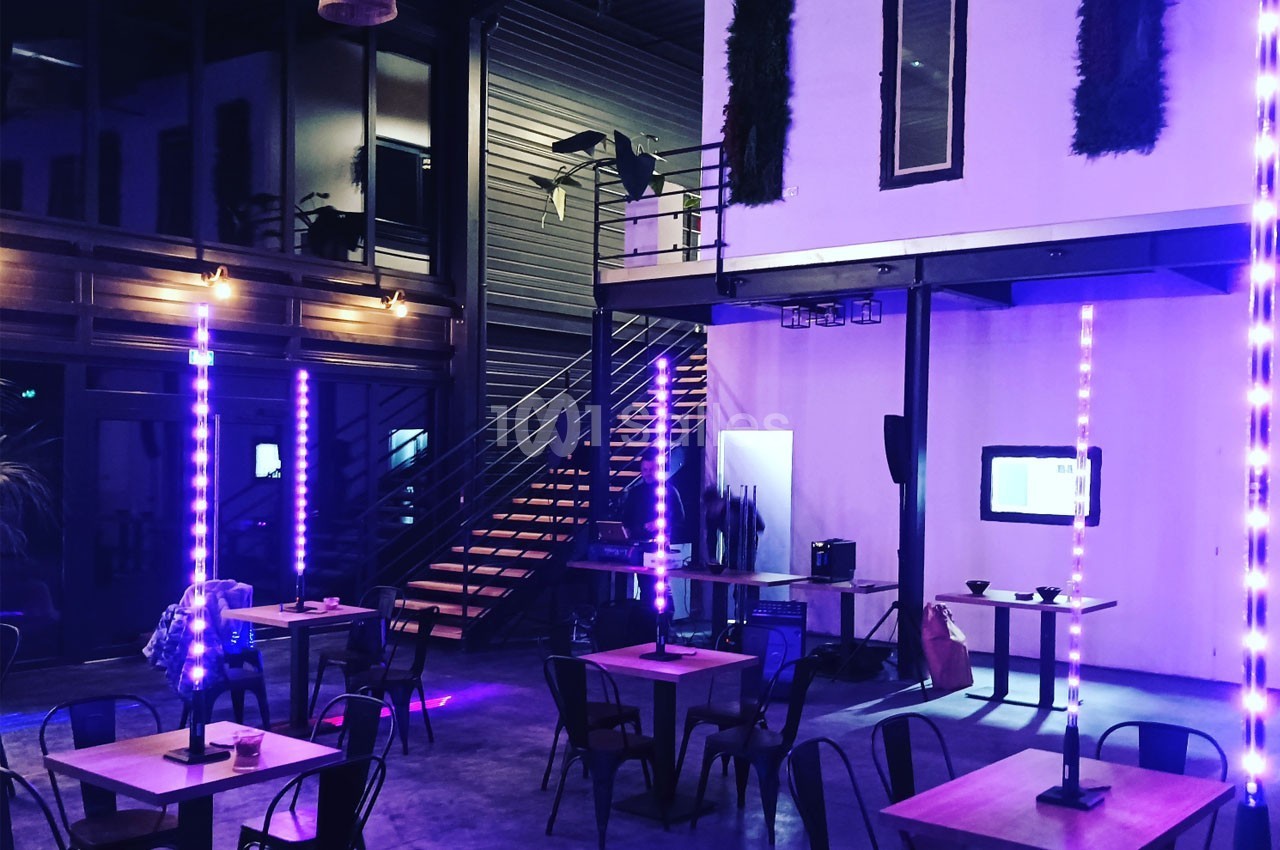 Salle moderne avec tables, chaises, éclairage violet et escalier métallique menant à une mezzanine.