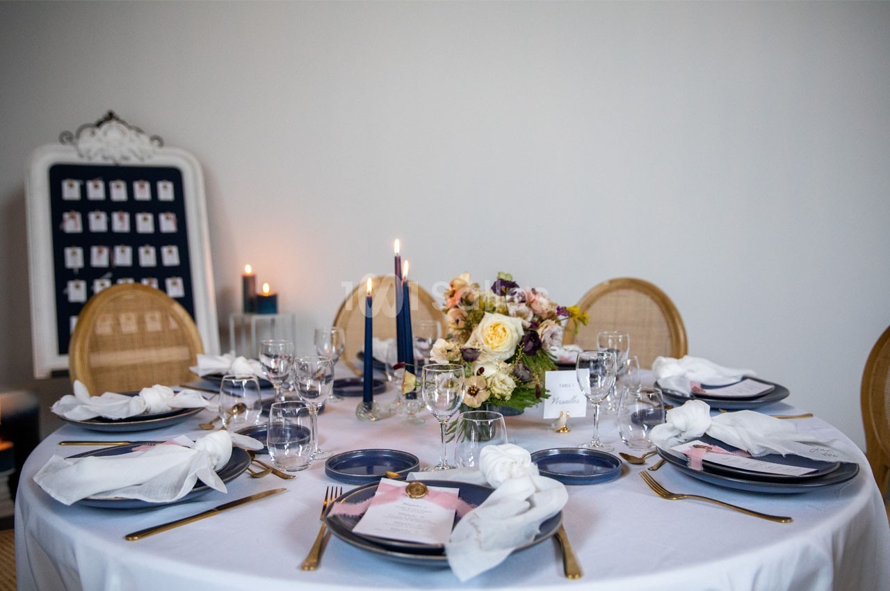 Table ronde élégamment dressée avec vaisselle bleue, serviettes blanches, bougies et bouquet de fleurs au centre.