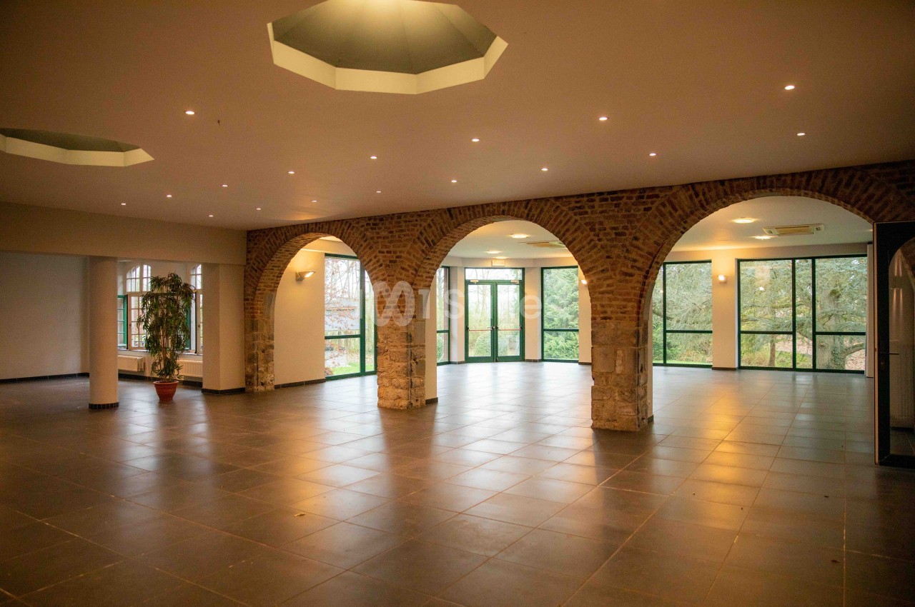 Salle spacieuse avec arches en briques, grandes fenêtres donnant sur un espace extérieur arboré et carrelage sombre.
