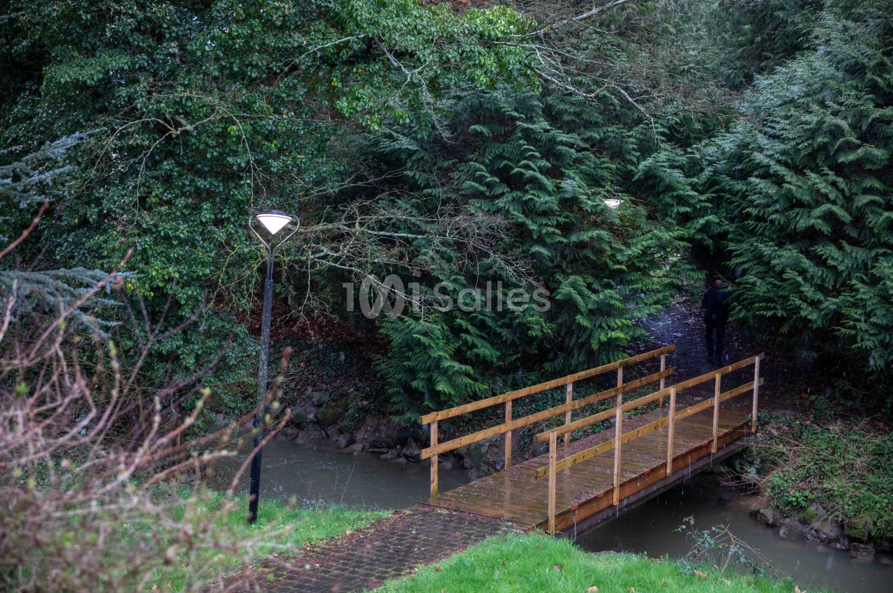 Passerelle en bois traversant un ruisseau, entourée de végétation dense et éclairée par des lampadaires.