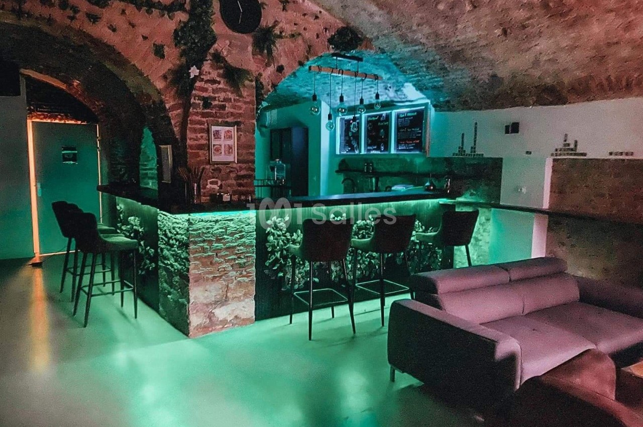 Bar avec comptoir en briques éclairé de lumières vertes, chaises hautes, canapé et voûtes en pierre apparente.