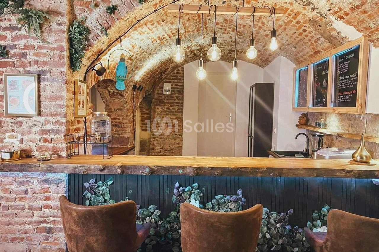 Bar avec comptoir en bois, tabourets rembourrés, murs en briques et plafond voûté éclairé par des ampoules suspendues.