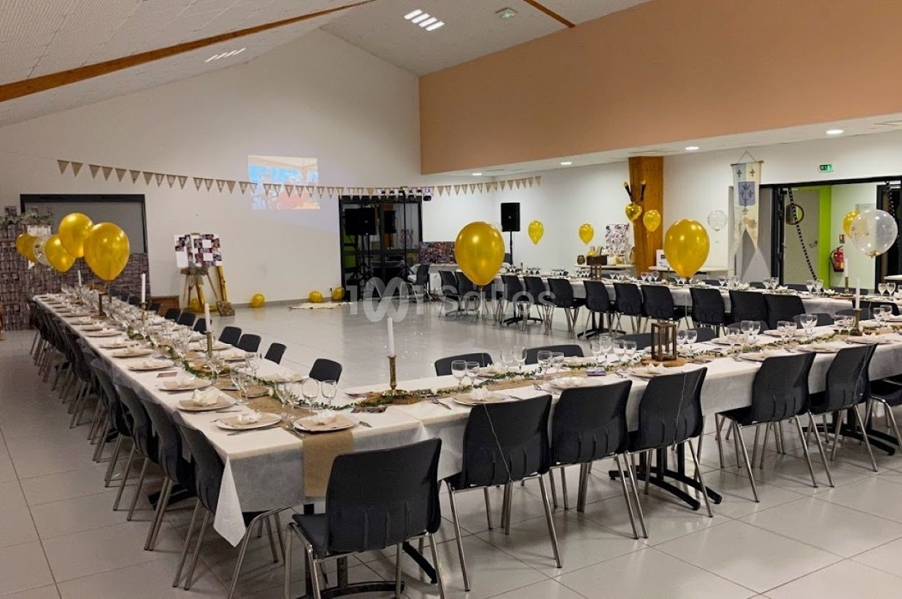 Salle décorée pour un événement, avec de longues tables dressées, des ballons dorés et des guirlandes en arrière-plan.