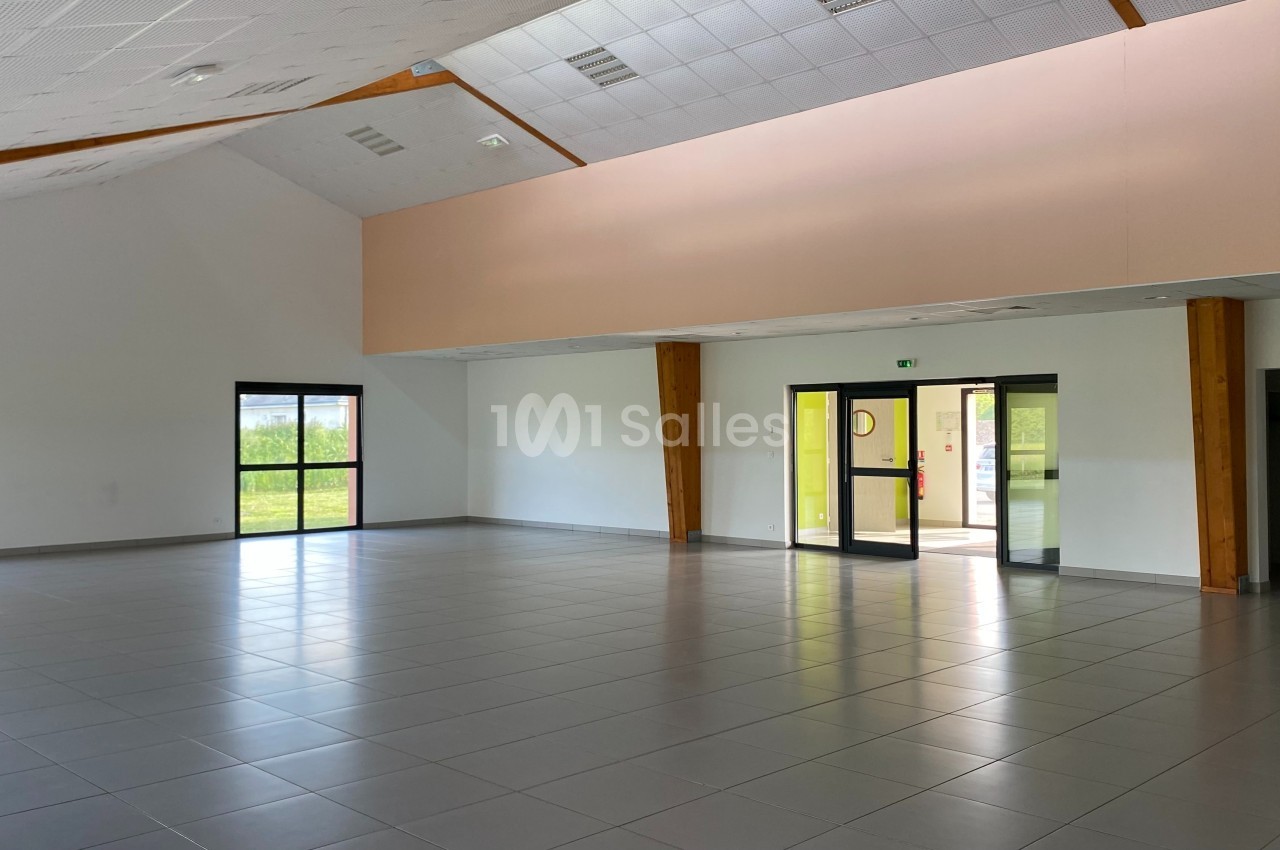 Salle vide avec carrelage gris, murs blancs, grandes fenêtres et portes vitrées laissant entrer la lumière naturelle.
