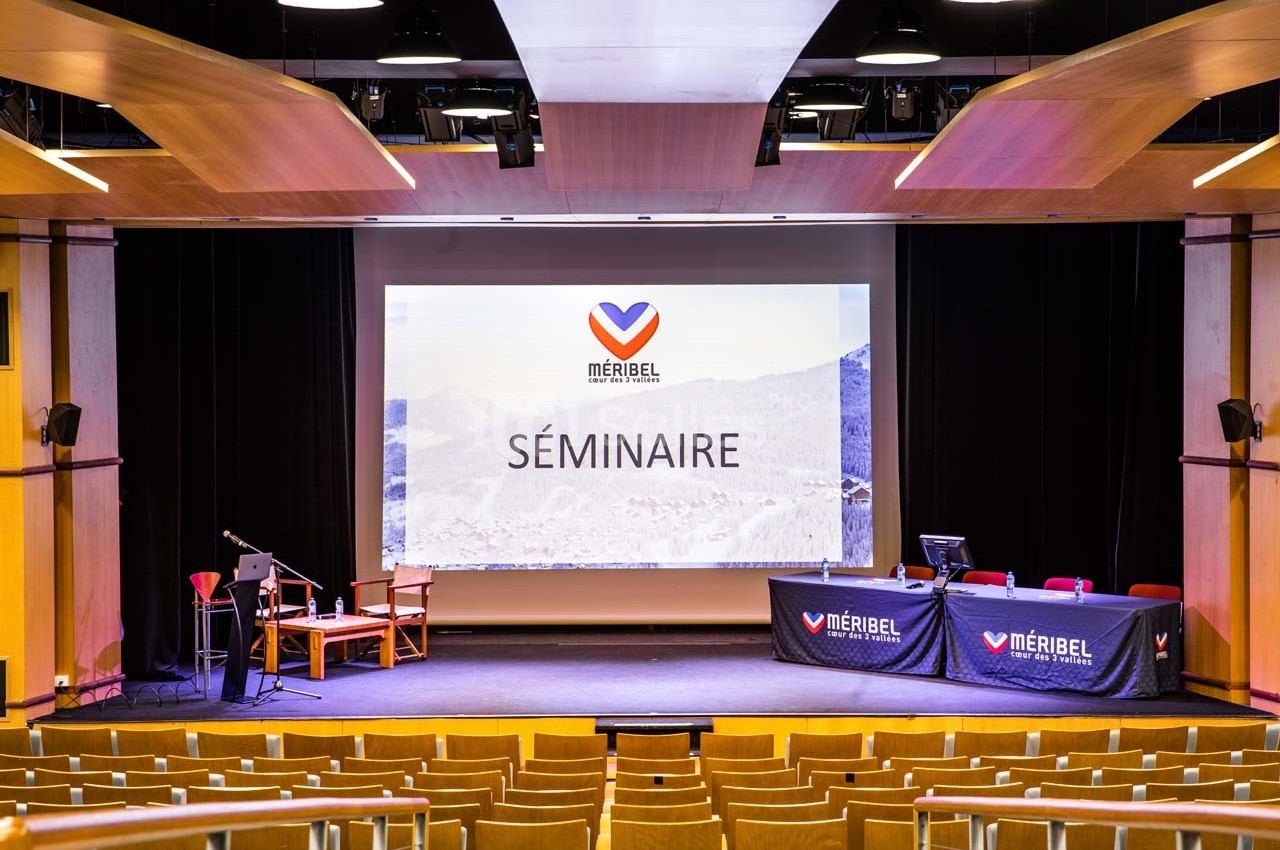 Salle de conférence avec scène, écran affichant ’Séminaire’ et tables équipées pour une présentation.