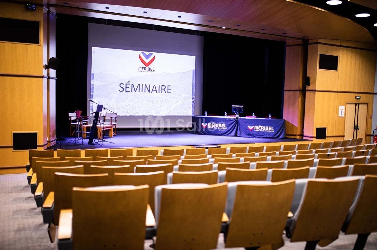 Salle de conférence vide avec des rangées de sièges en bois, un écran affichant ’Séminaire’ et une estrade équipée.