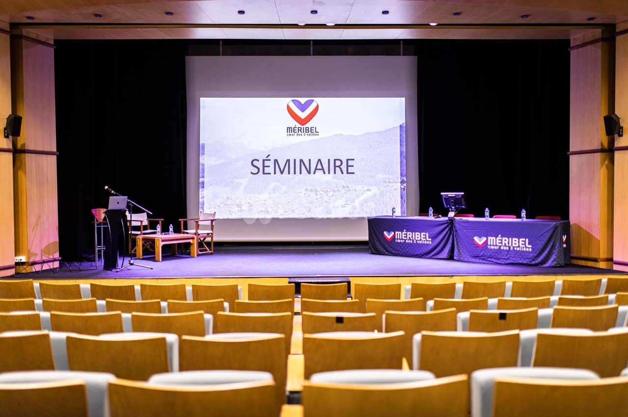 Salle de conférence avec écran de projection affichant ’Séminaire’, chaises alignées et table équipée sur scène.