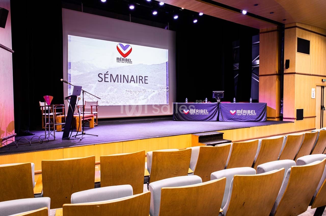Salle de conférence avec écran de projection affichant ’Séminaire’, deux tables et des chaises sur scène.