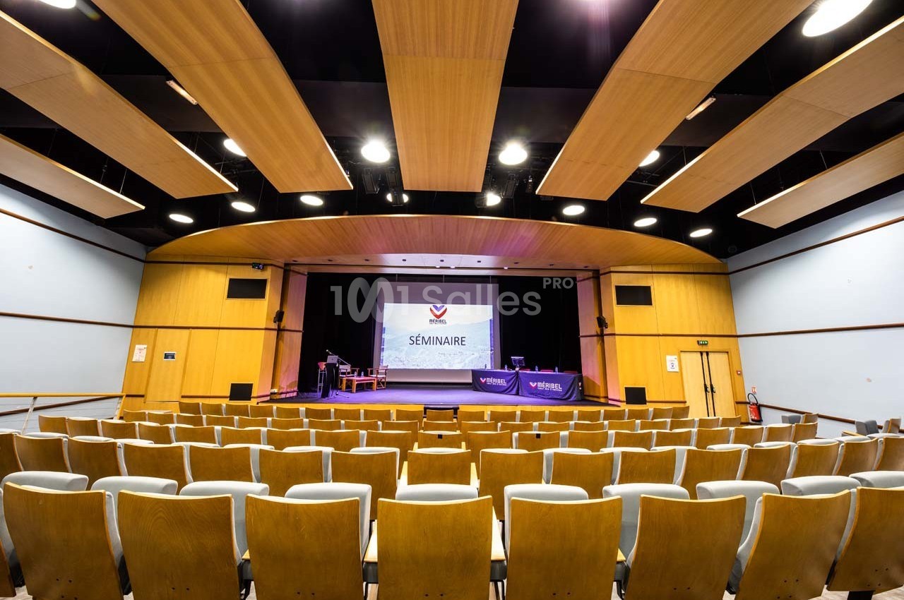 Salle de conférence vide avec des rangées de sièges, une scène équipée et un écran affichant ’Séminaire’.