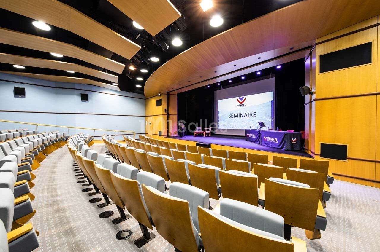 Salle de conférence vide avec des rangées de sièges, une scène équipée et un écran affichant le mot ’Séminaire’.