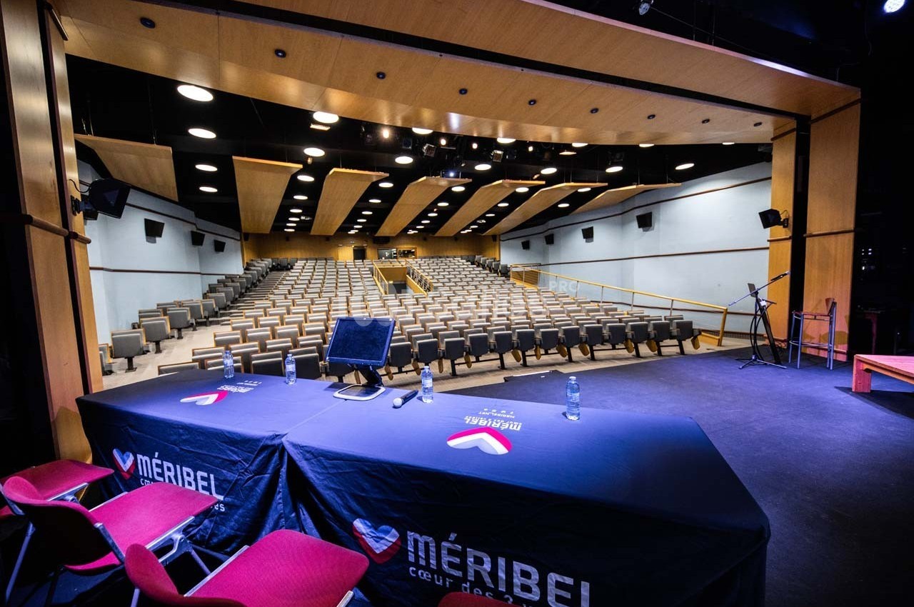 Salle de conférence moderne avec rangées de sièges, vue depuis une scène équipée de micros et d'une table.