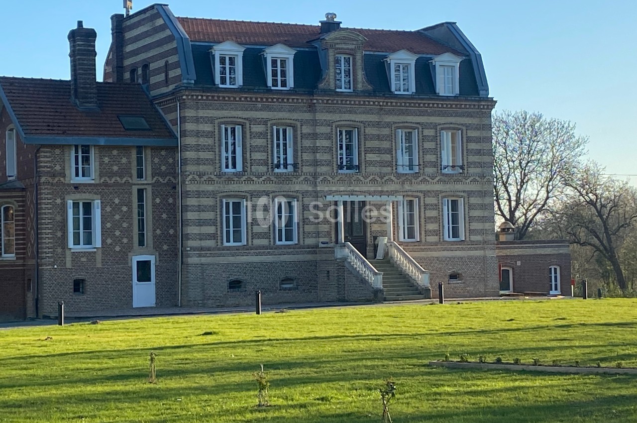 Façade d'un manoir en briques avec un toit mansardé, entouré d'une pelouse et d'arbres en arrière-plan.