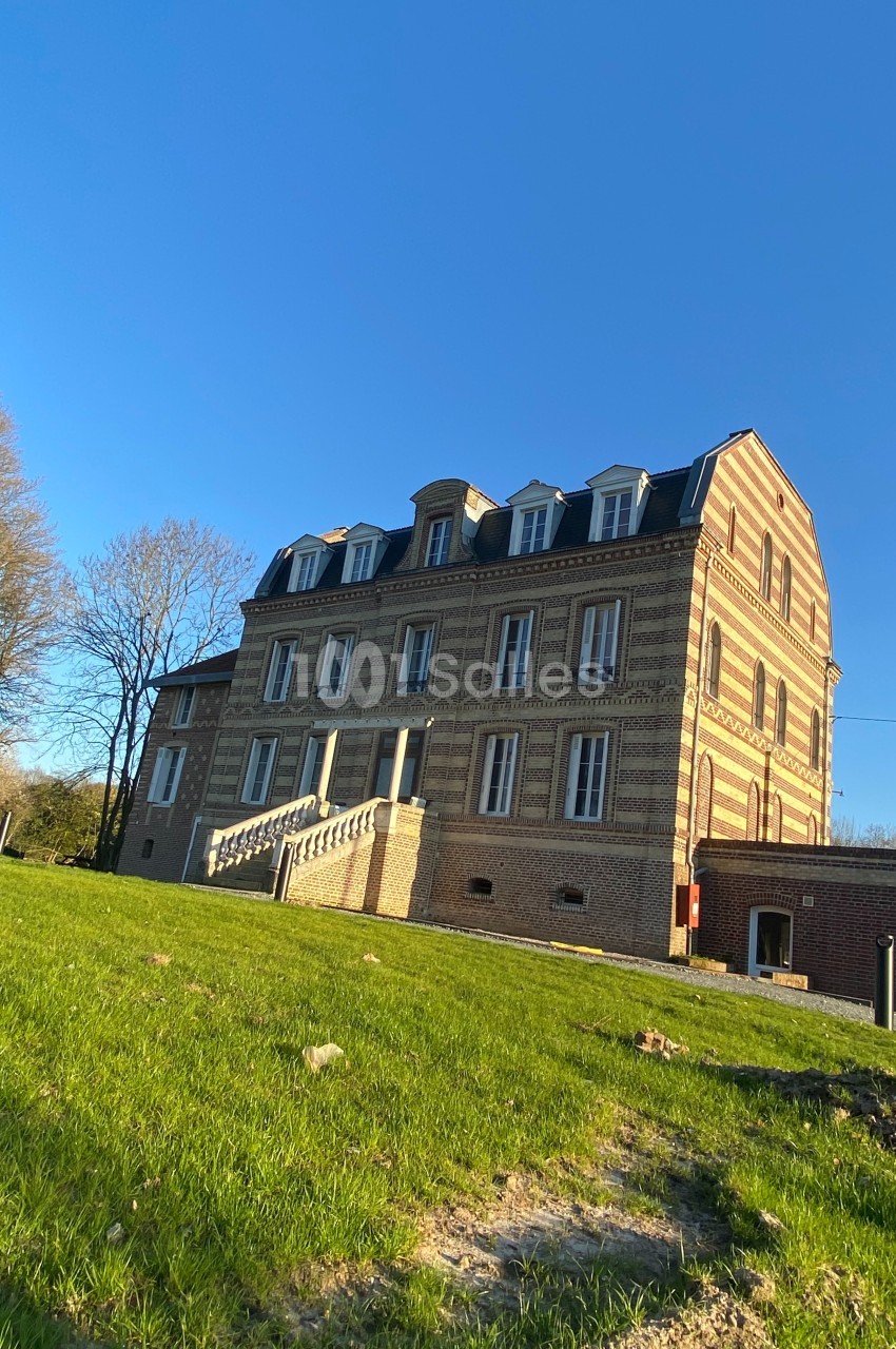 Bâtiment en briques à trois étages avec toit mansardé, entouré de pelouse et d'arbres sous un ciel bleu.