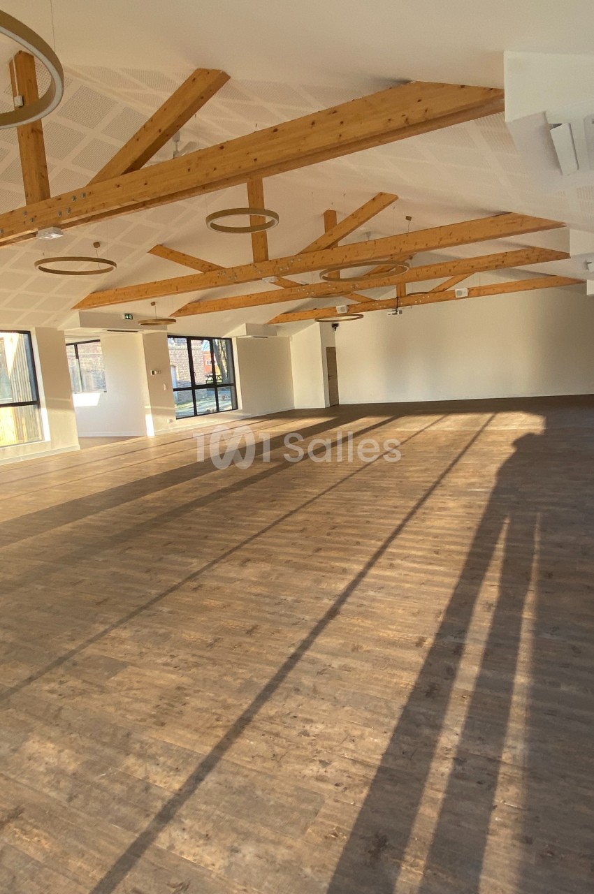 Salle lumineuse avec parquet en bois, poutres apparentes au plafond et grandes fenêtres laissant entrer la lumière naturelle.