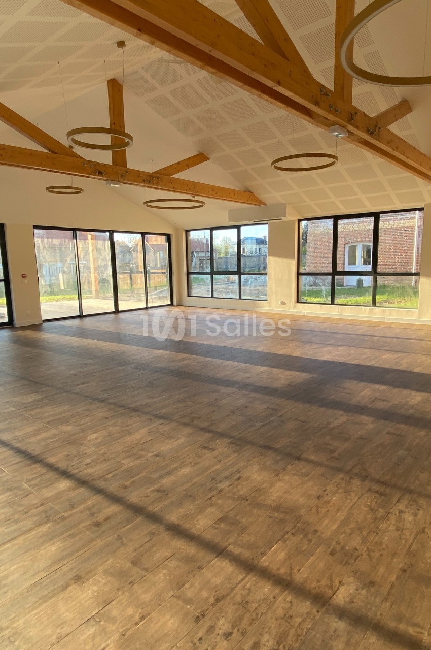 Salle vide avec parquet en bois, grandes baies vitrées laissant entrer la lumière naturelle et poutres apparentes au plafond.