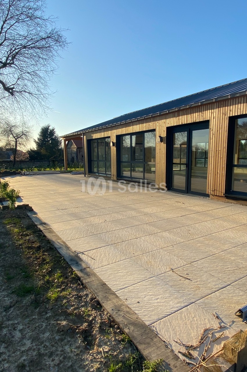 Terrasse en dalles claires devant une maison moderne en bois avec grandes baies vitrées, sous un ciel dégagé.