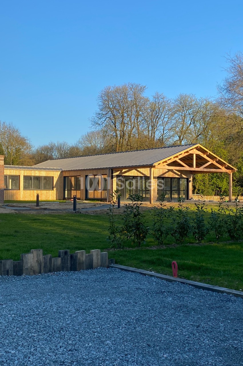 Bâtiment moderne en bois avec de grandes baies vitrées, entouré de pelouse et d'arbres sous un ciel dégagé.