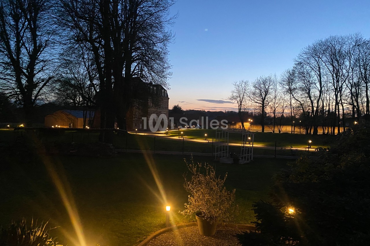 Jardin éclairé par des lampes au sol au crépuscule, avec des arbres et un bâtiment en arrière-plan.