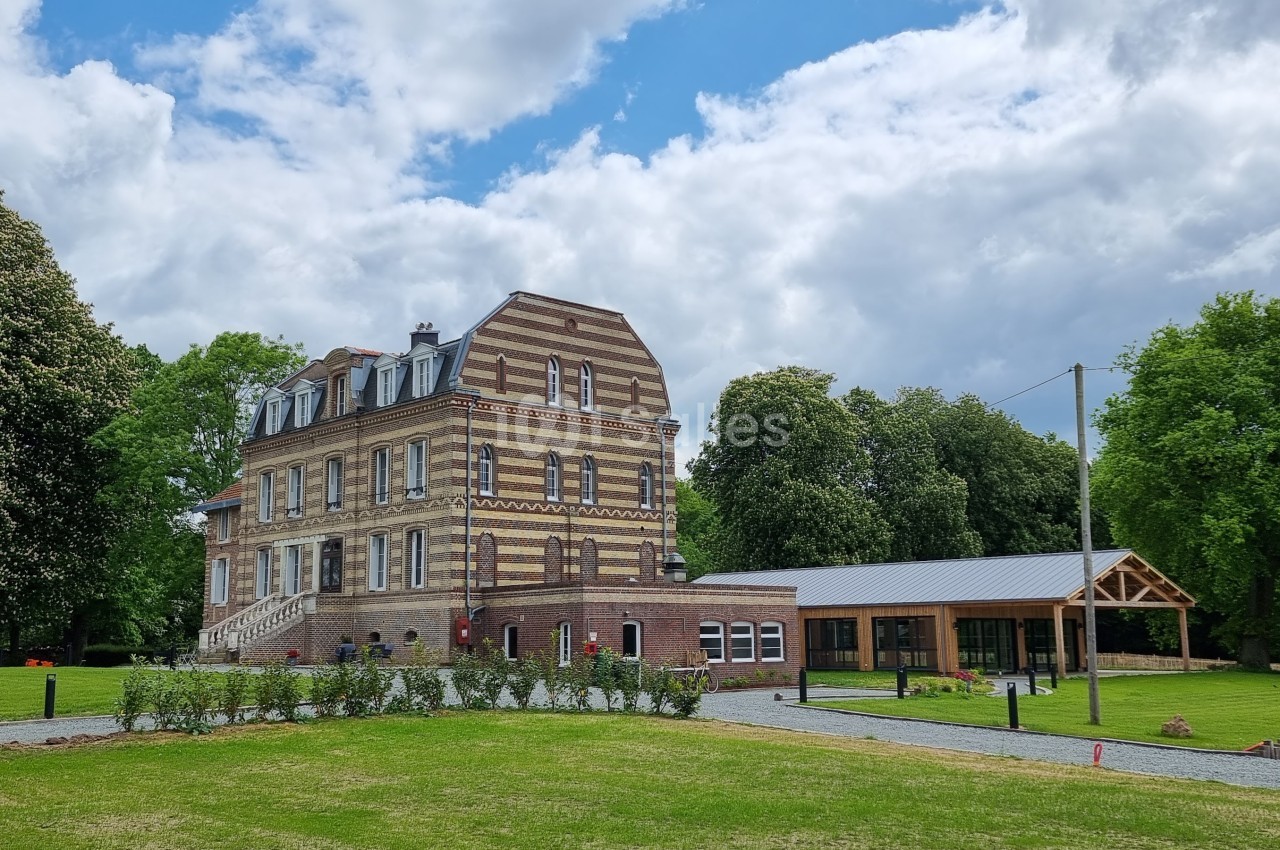 Grand bâtiment en briques entouré d'arbres, avec une extension moderne et une pelouse bien entretenue au premier plan.