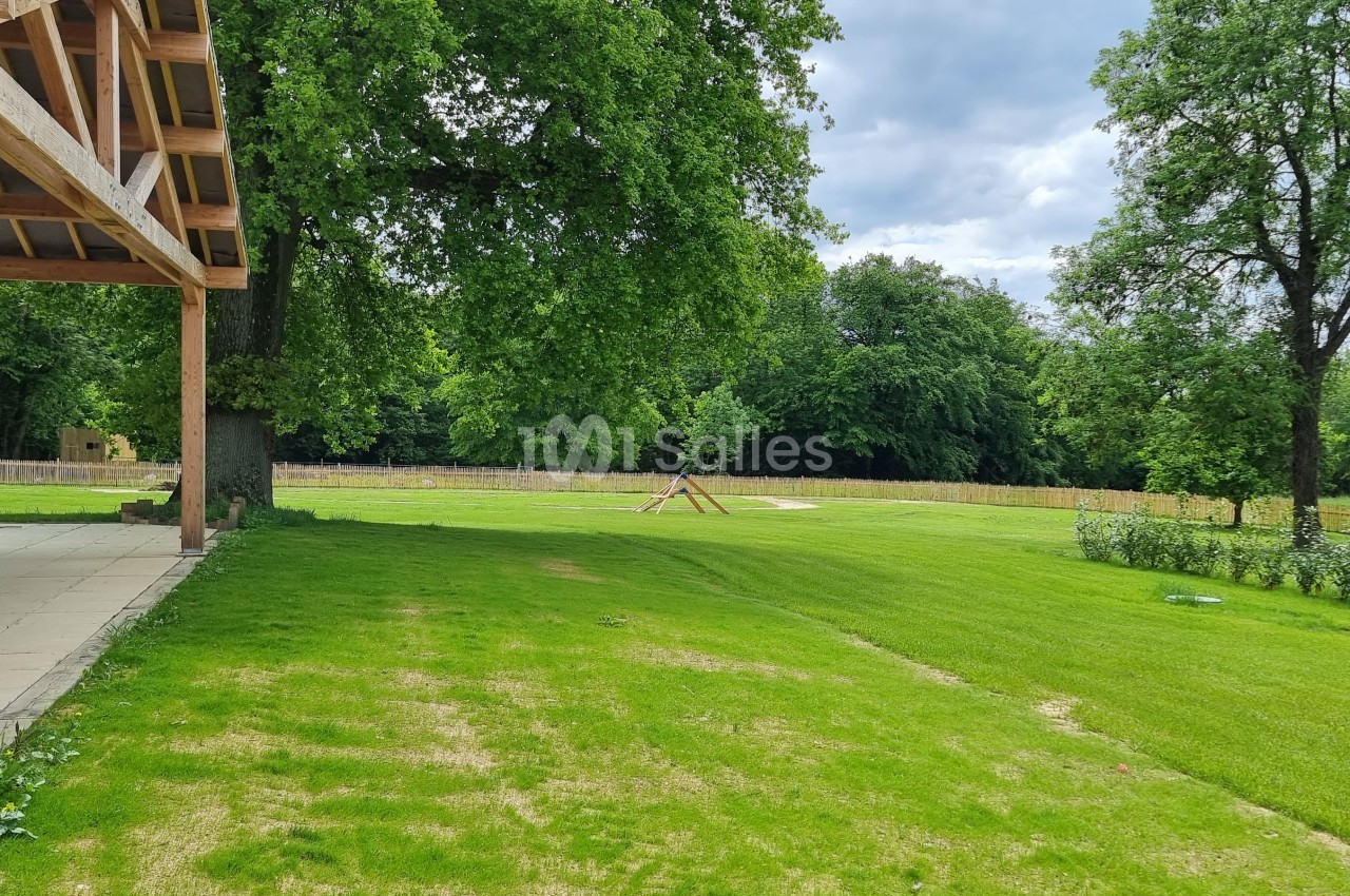 Vue d'un jardin verdoyant avec pelouse, arbres et une structure en bois sur la gauche sous un ciel partiellement nuageux.
