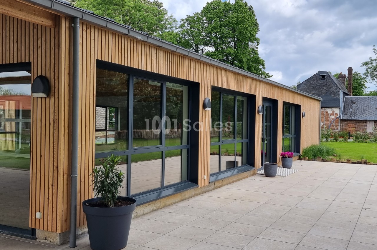 Bâtiment moderne avec façade en bois, grandes baies vitrées et terrasse pavée, entouré d'un jardin verdoyant.