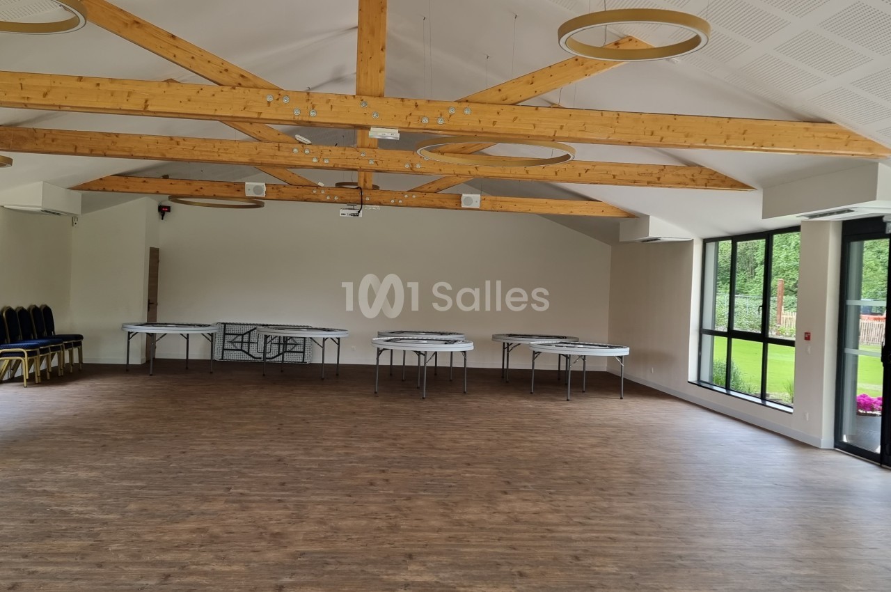 Salle lumineuse avec parquet, poutres apparentes, grandes baies vitrées et tables pliées contre les murs.