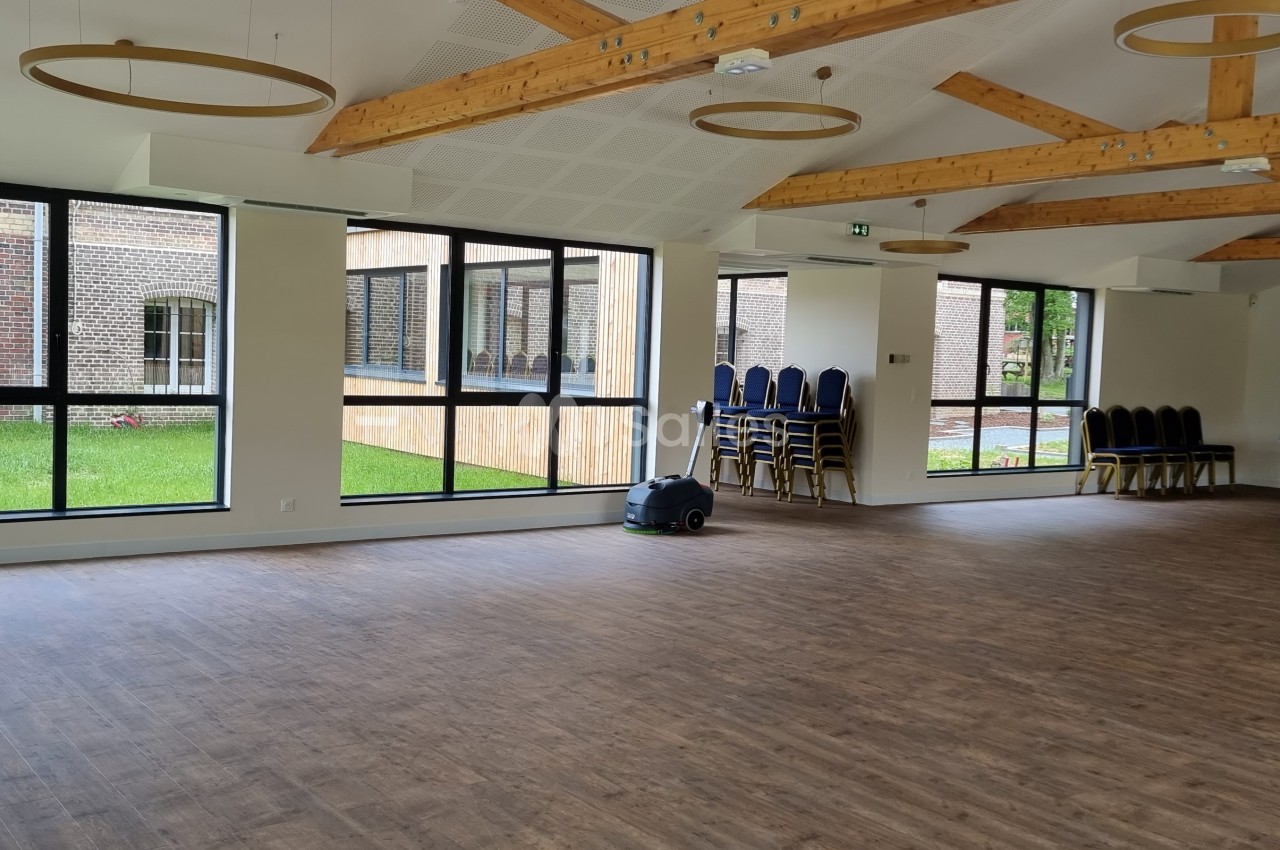 Salle lumineuse avec parquet, poutres apparentes, grandes fenêtres donnant sur un espace vert, chaises empilées sur le côté.