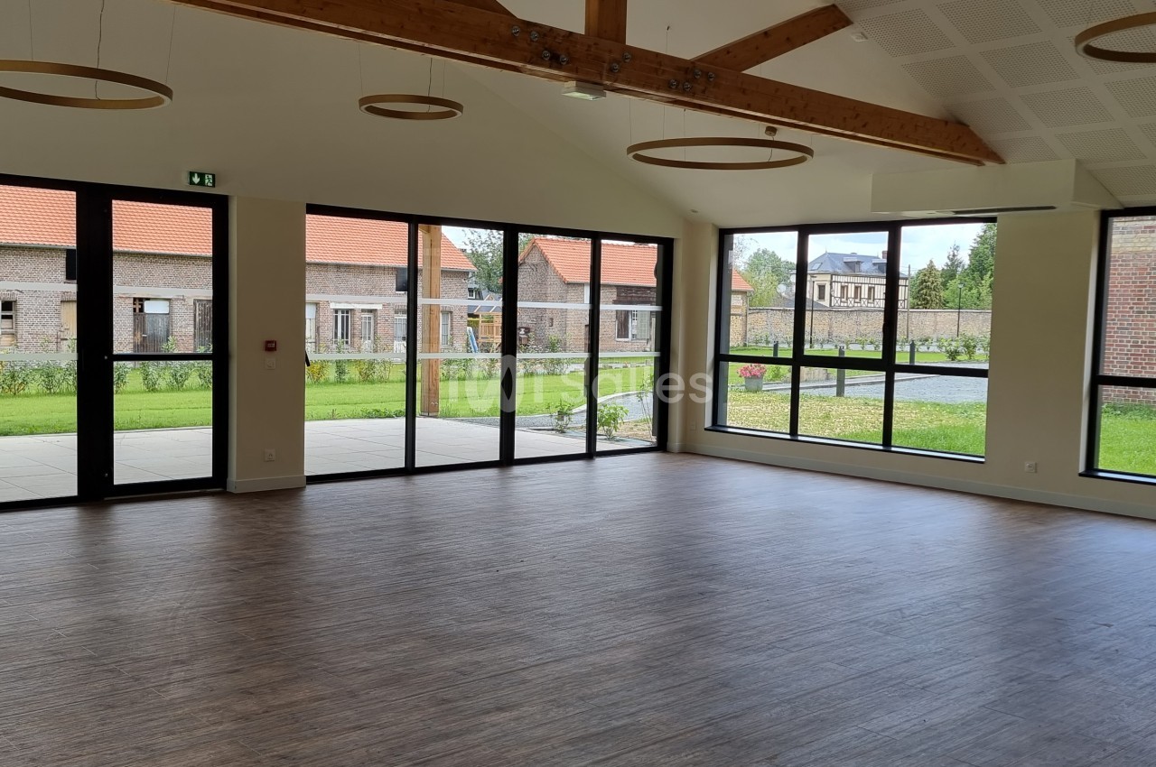 Salle lumineuse avec parquet, grandes baies vitrées donnant sur un jardin et poutres apparentes au plafond.