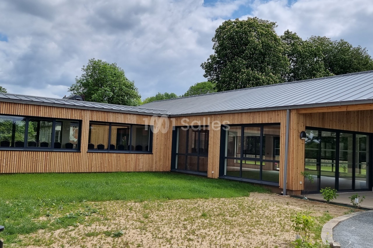 Bâtiment moderne en bois avec de grandes baies vitrées, entouré de pelouse et d'arbres sous un ciel partiellement nuageux.