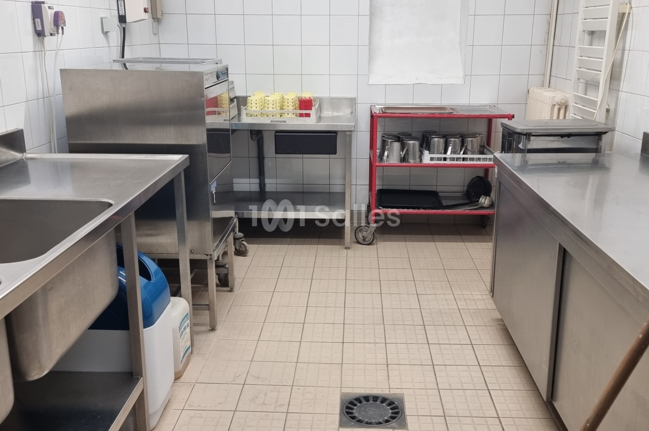 Cuisine professionnelle avec équipements en inox, évier double, étagères, carrelage blanc et sol en carrelage beige.