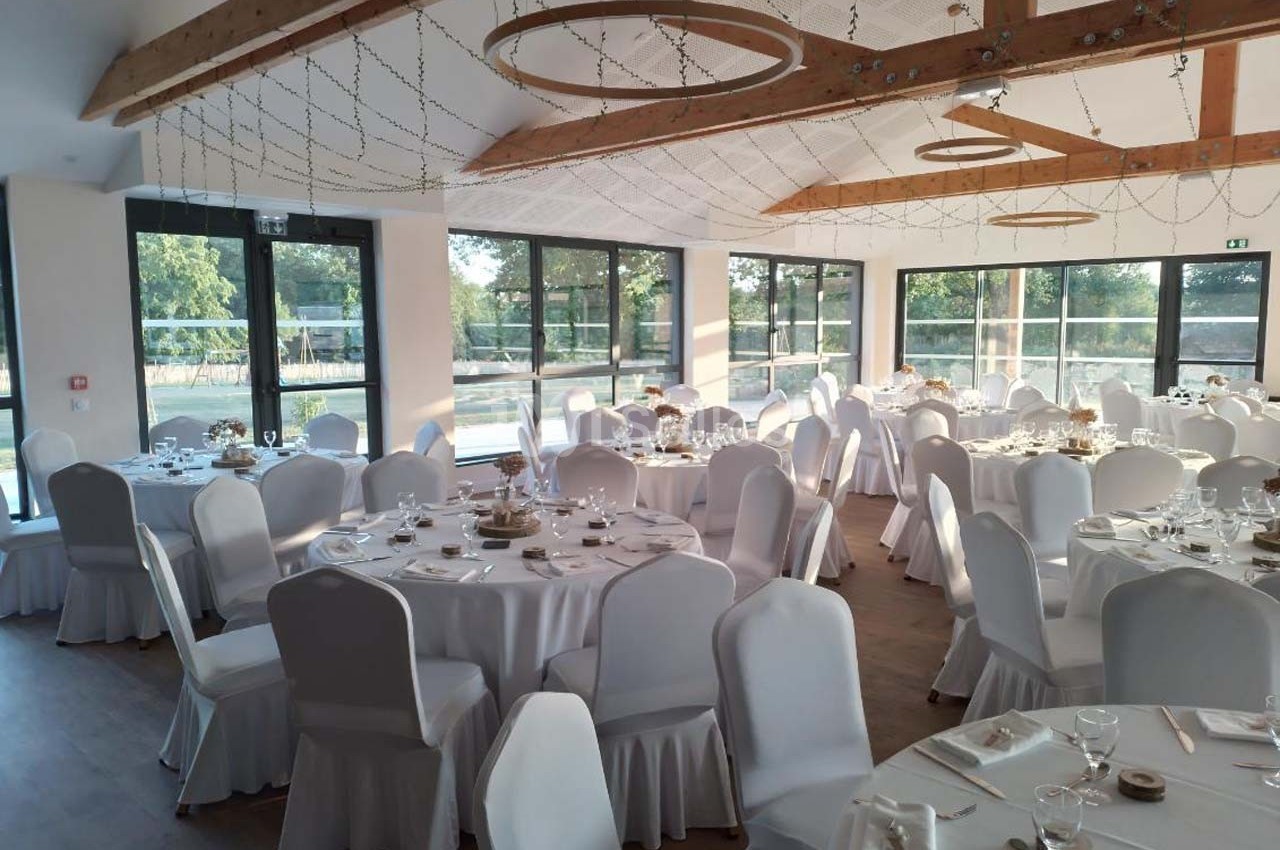 Salle de réception lumineuse avec tables rondes dressées, nappes blanches et grandes baies vitrées donnant sur l'extérieur.