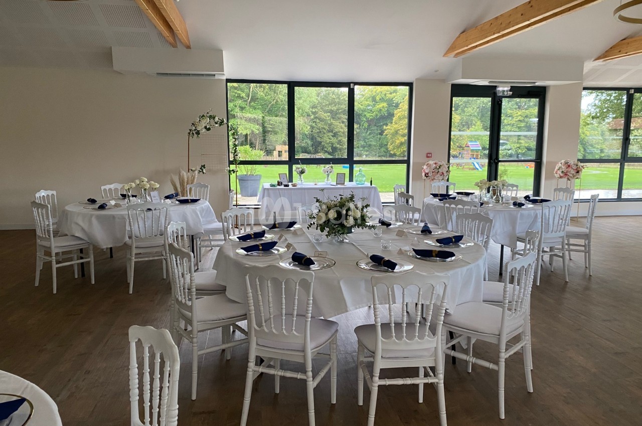 Salle de réception lumineuse avec tables rondes dressées, chaises blanches et vue sur un jardin verdoyant.