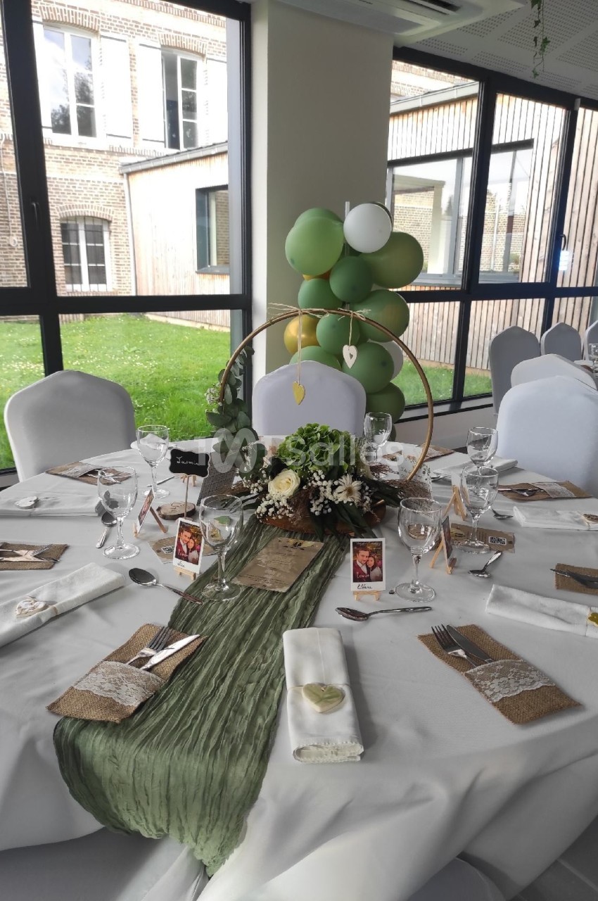 Table de réception décorée avec nappes blanches, chemin de table vert, compositions florales et ballons verts en arrière…