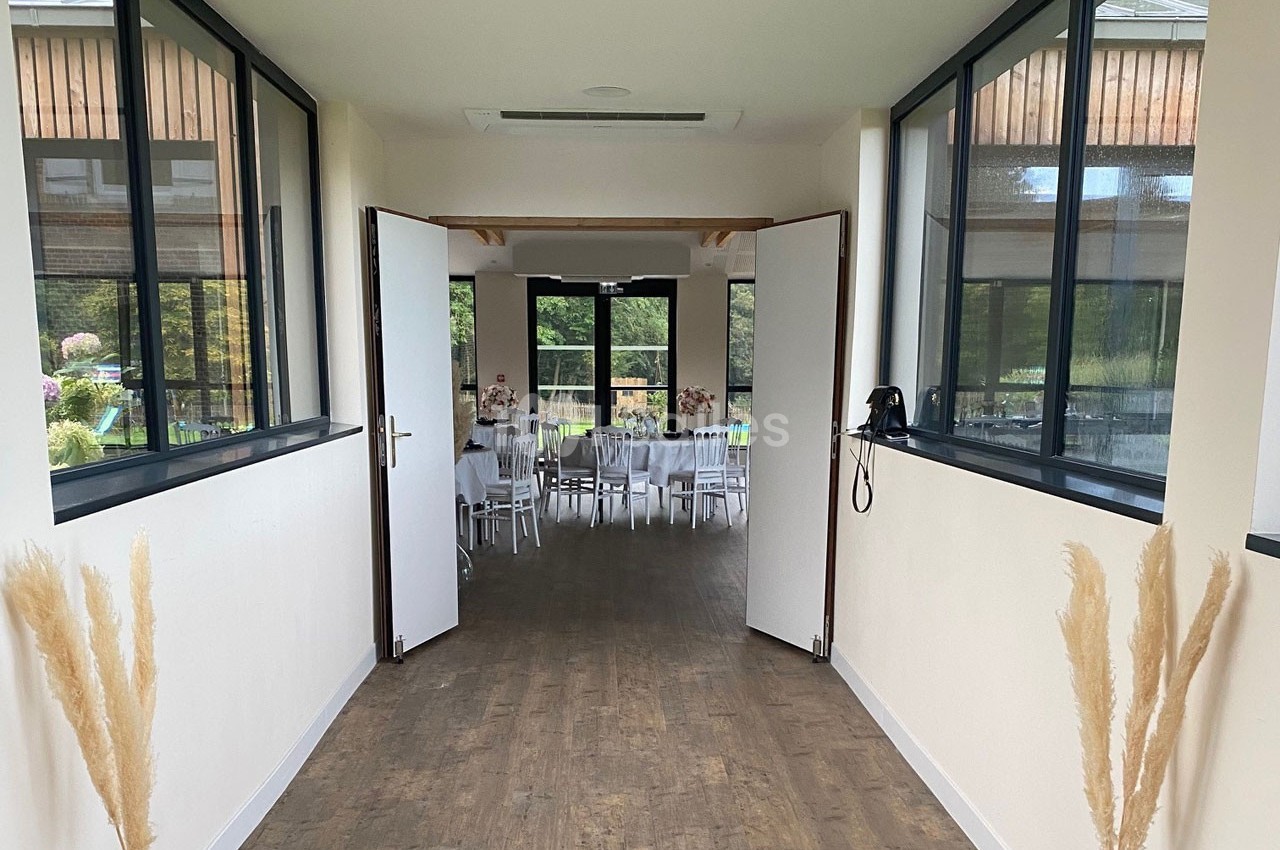 Vue d'un couloir lumineux menant à une salle de réception avec des tables dressées et des baies vitrées donnant sur un…