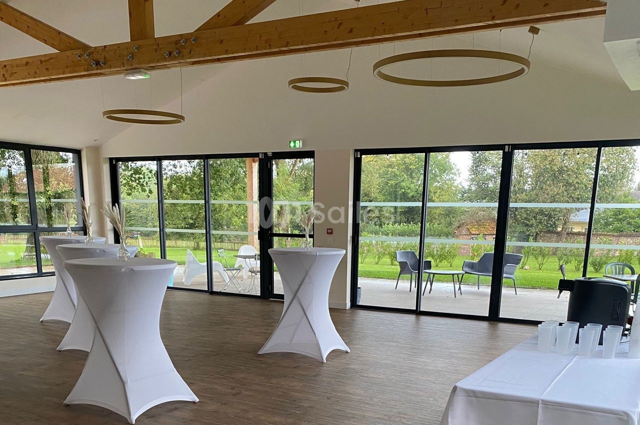 Salle lumineuse avec grandes baies vitrées, tables hautes nappées de blanc et vue sur un espace extérieur verdoyant.