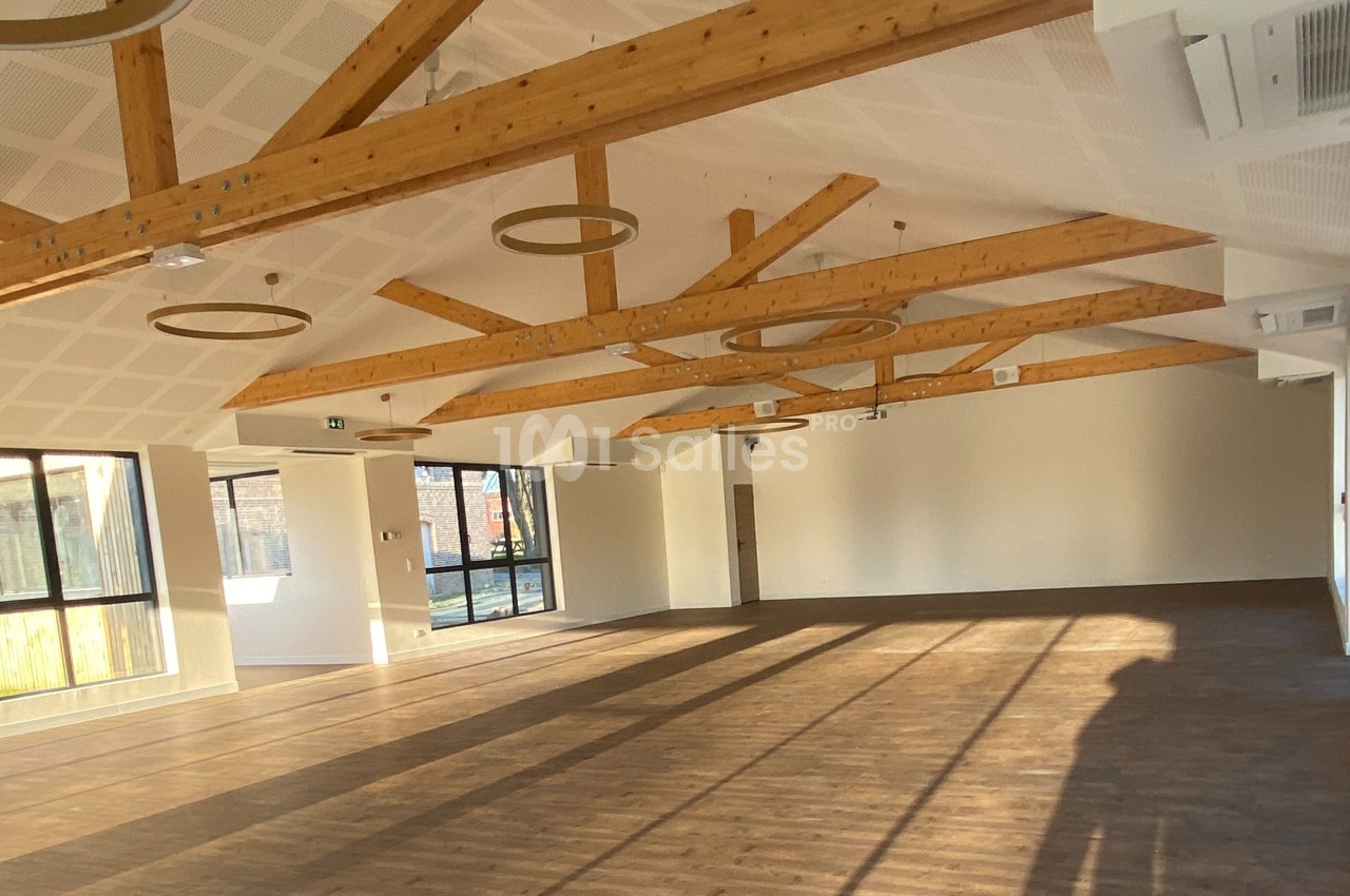 Salle lumineuse avec parquet en bois, poutres apparentes au plafond et grandes fenêtres laissant entrer la lumière naturelle.