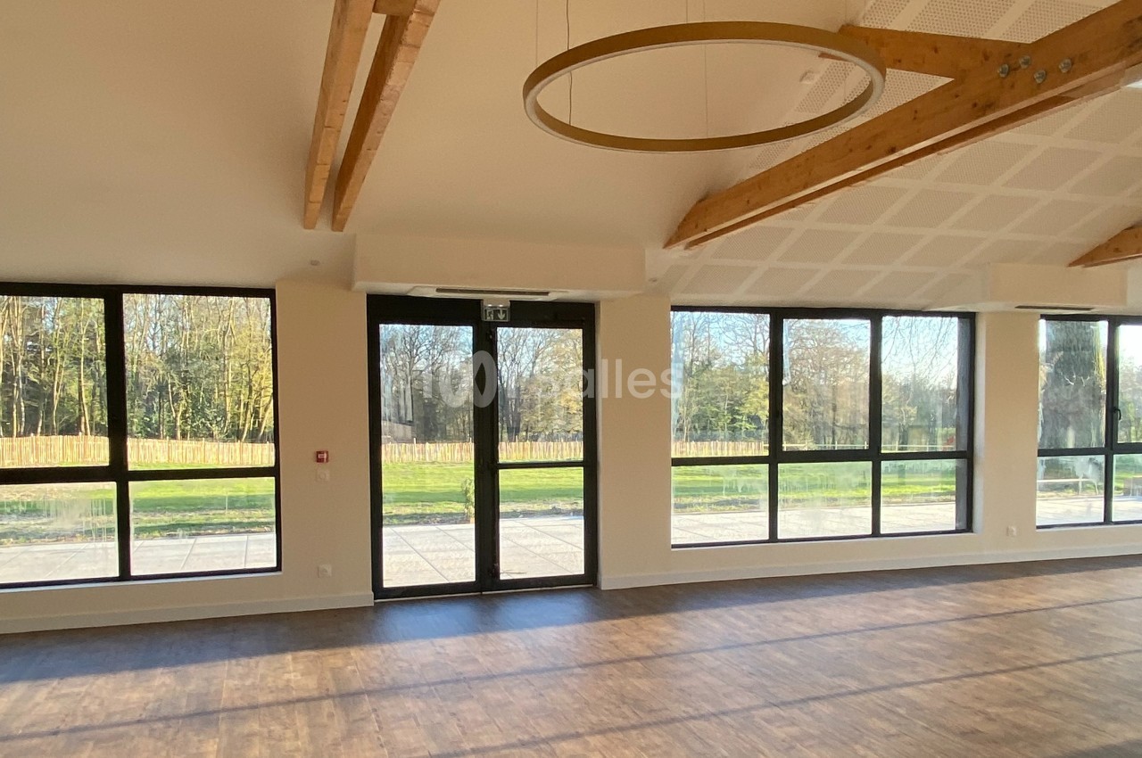 Grande salle lumineuse avec parquet, poutres apparentes et larges baies vitrées donnant sur un espace extérieur arboré.