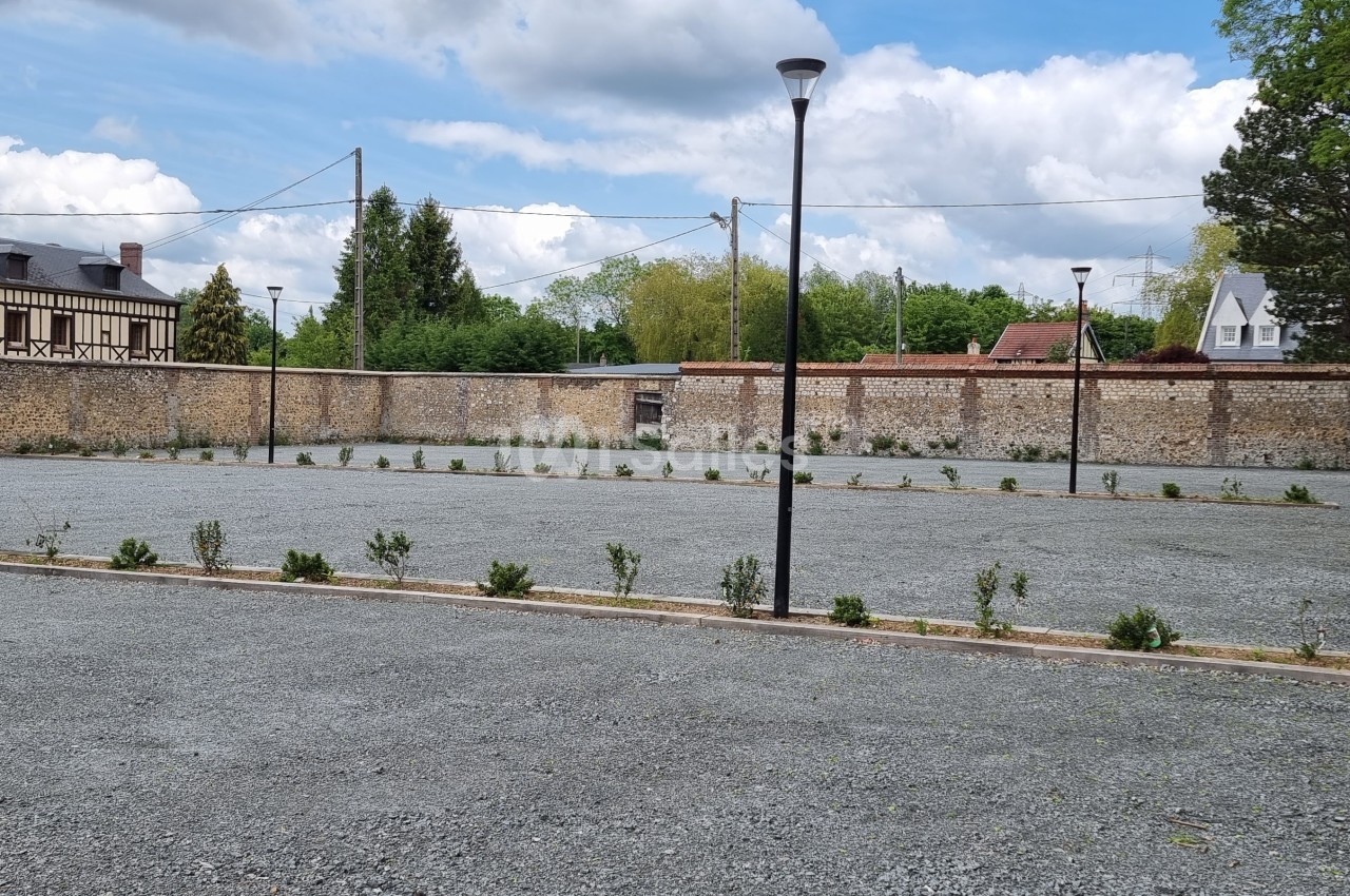 Parking en gravier avec quelques jeunes arbustes, entouré de murs en pierre et bordé de maisons et d'arbres.