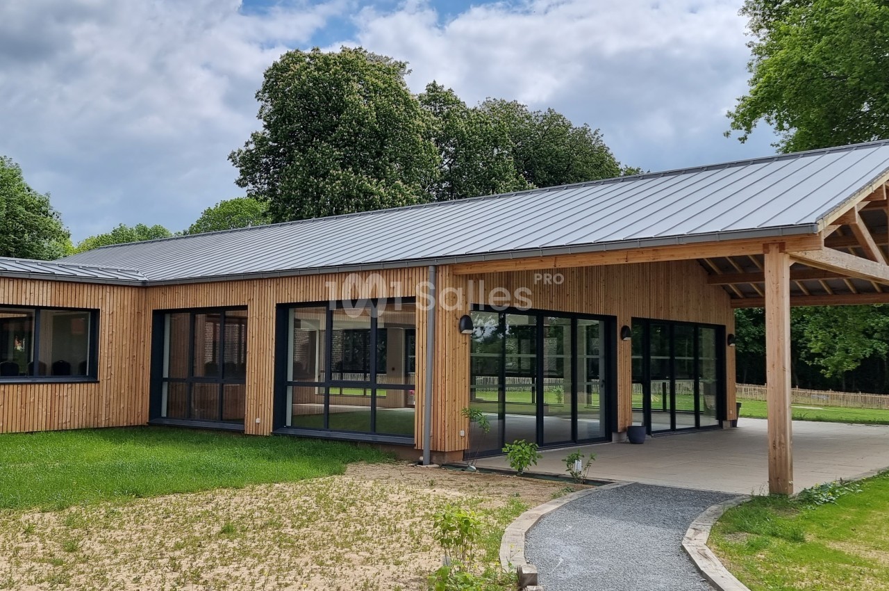 Bâtiment moderne en bois avec de grandes baies vitrées, entouré de pelouse et d'arbres sous un ciel partiellement nuageux.