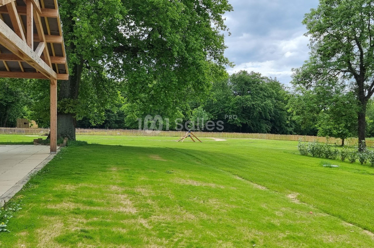 Vue d'un grand jardin verdoyant avec pelouse, arbres et une structure en bois sur la gauche.