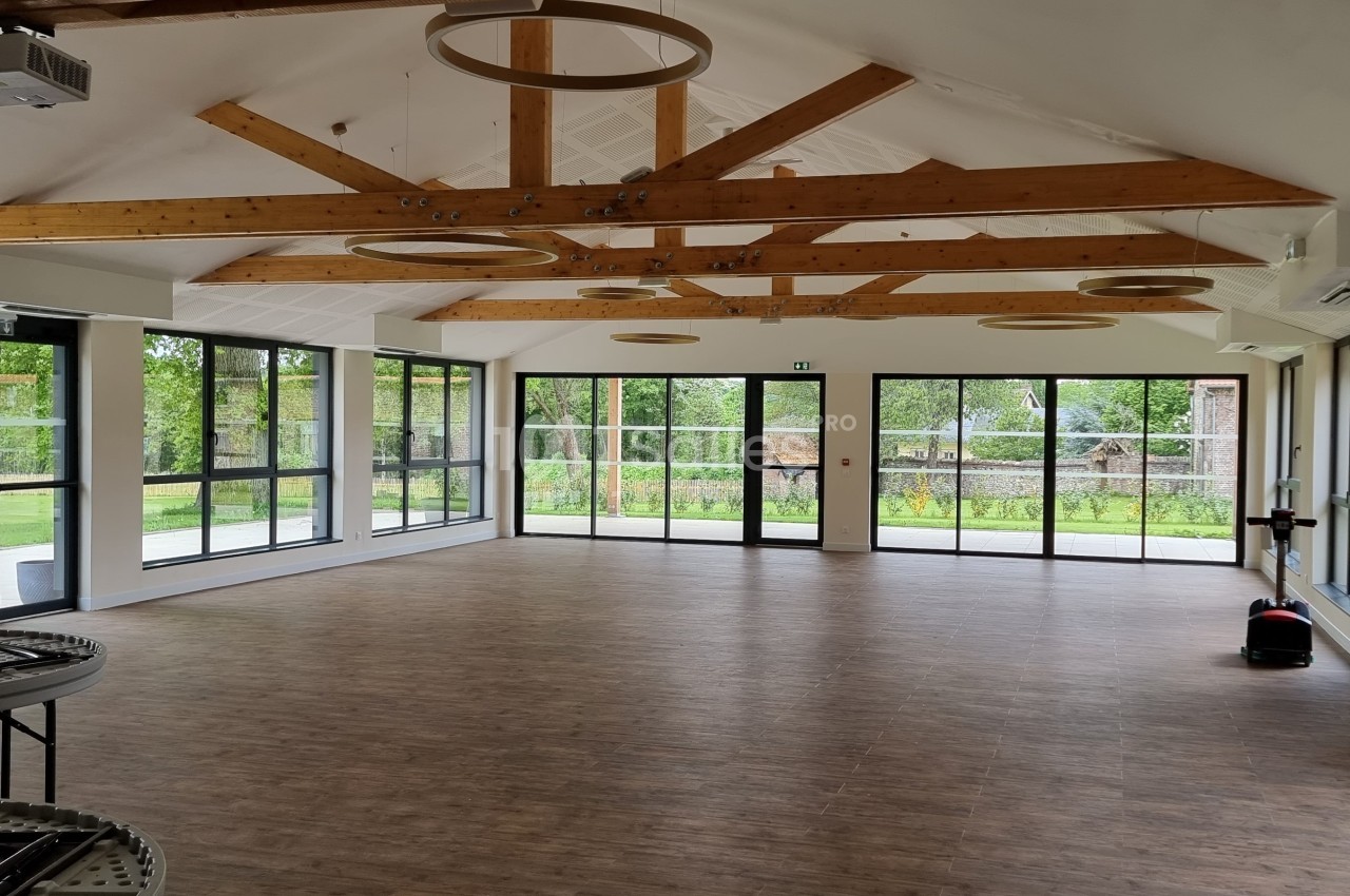 Salle lumineuse avec parquet, poutres apparentes, grandes baies vitrées donnant sur un espace extérieur verdoyant.