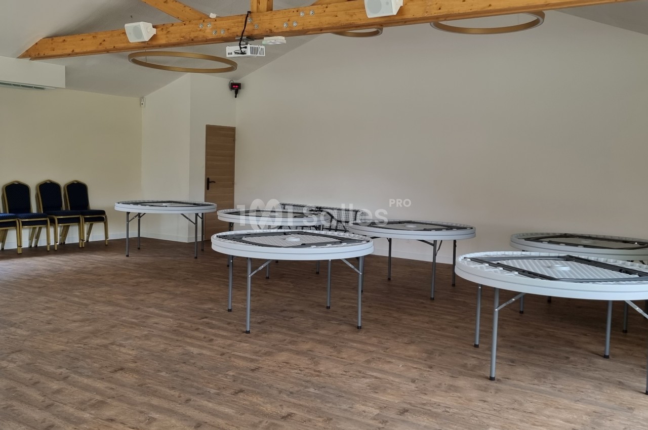 Salle lumineuse avec parquet, tables pliantes rondes disposées et chaises empilées près d'une fenêtre.