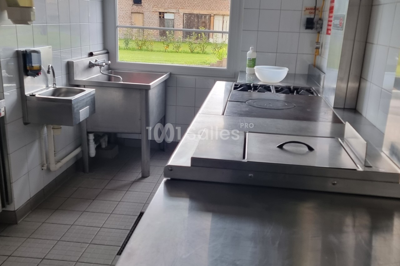 Cuisine professionnelle avec plan de travail en inox, évier, plaques de cuisson et fenêtre donnant sur un extérieur.