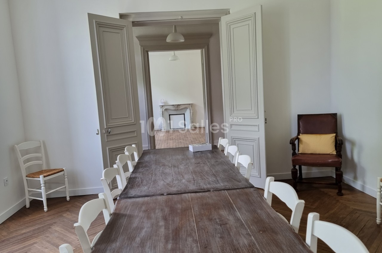 Salle lumineuse avec une grande table en bois entourée de chaises blanches, une cheminée visible en arrière-plan.