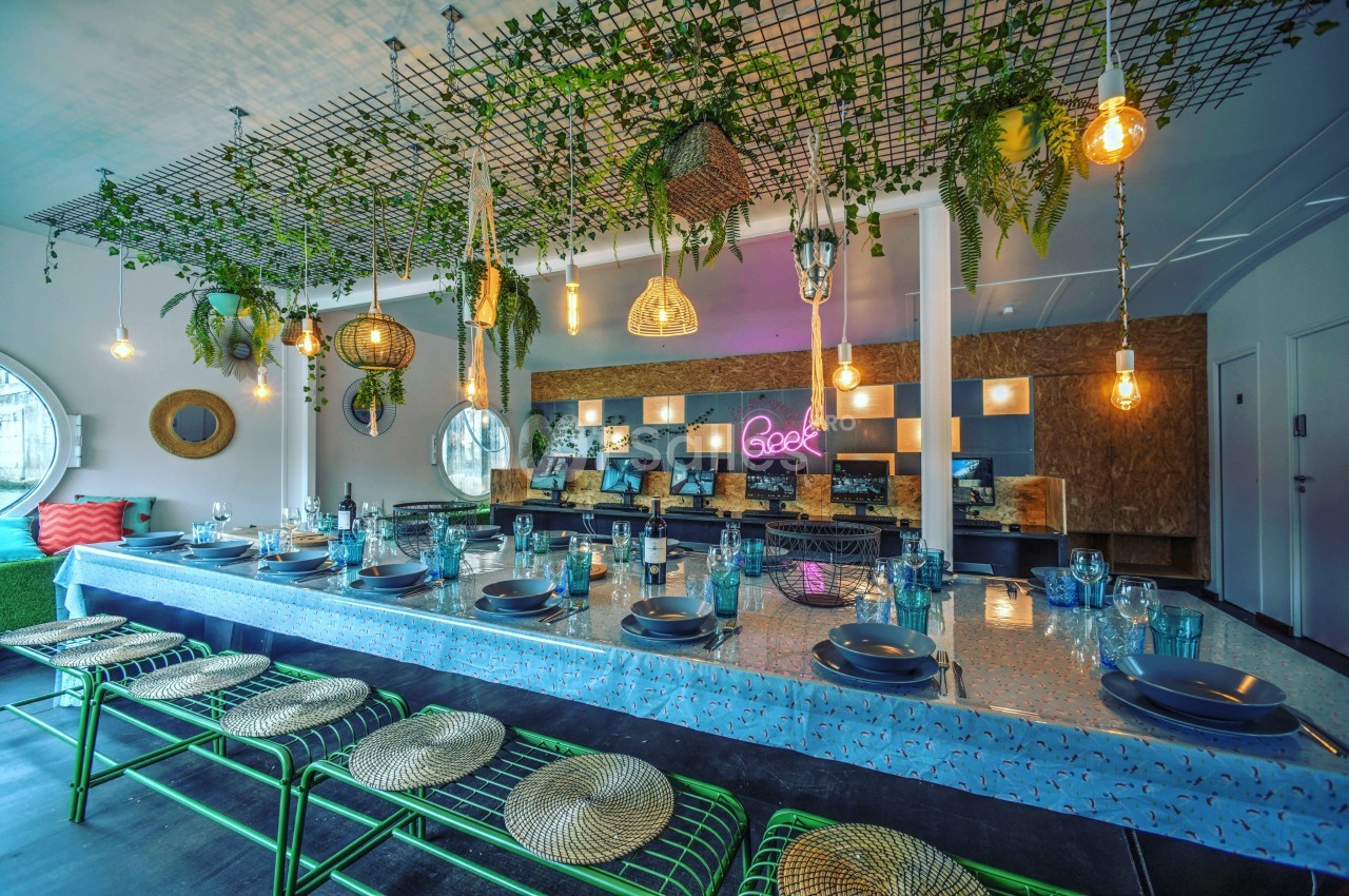 Grande table décorée avec vaisselle, entourée de chaises, sous un plafond orné de plantes suspendues et luminaires.