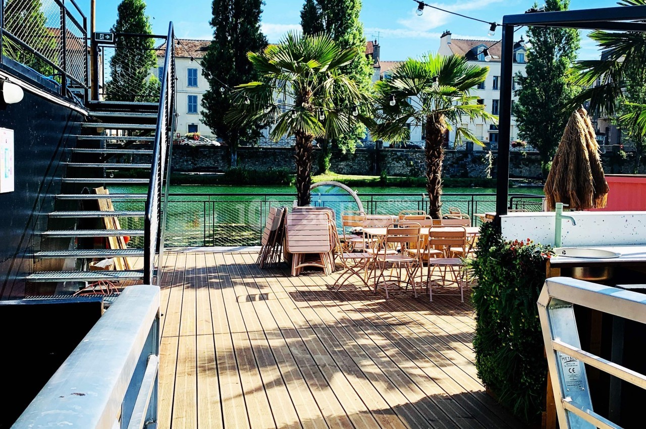 Terrasse en bois avec tables et chaises, bordée de palmiers, donnant sur un canal et des bâtiments en arrière-plan.
