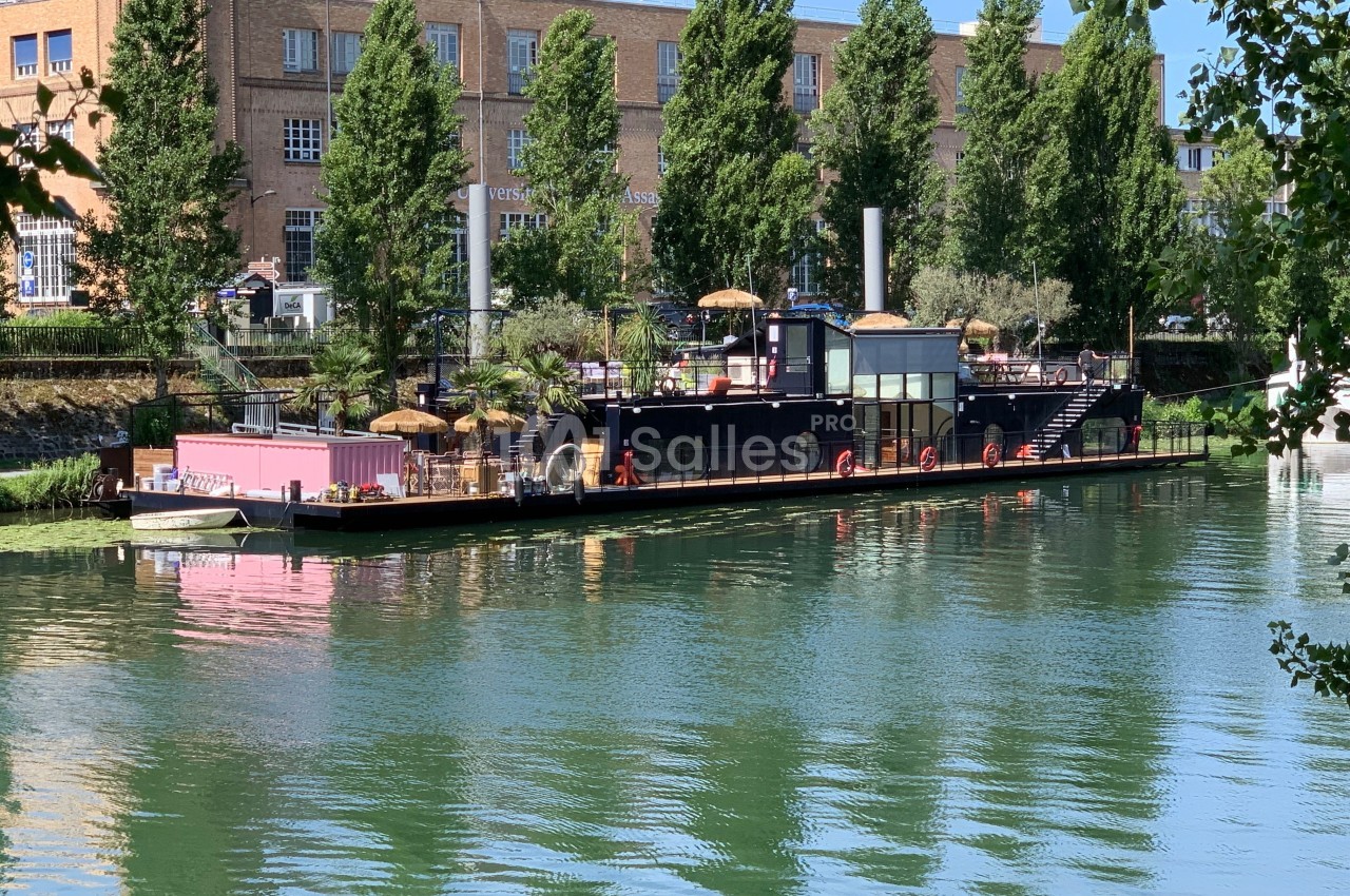 Bateau aménagé en terrasse flottante avec parasols, amarré sur une rivière bordée d'arbres et de bâtiments en briques.