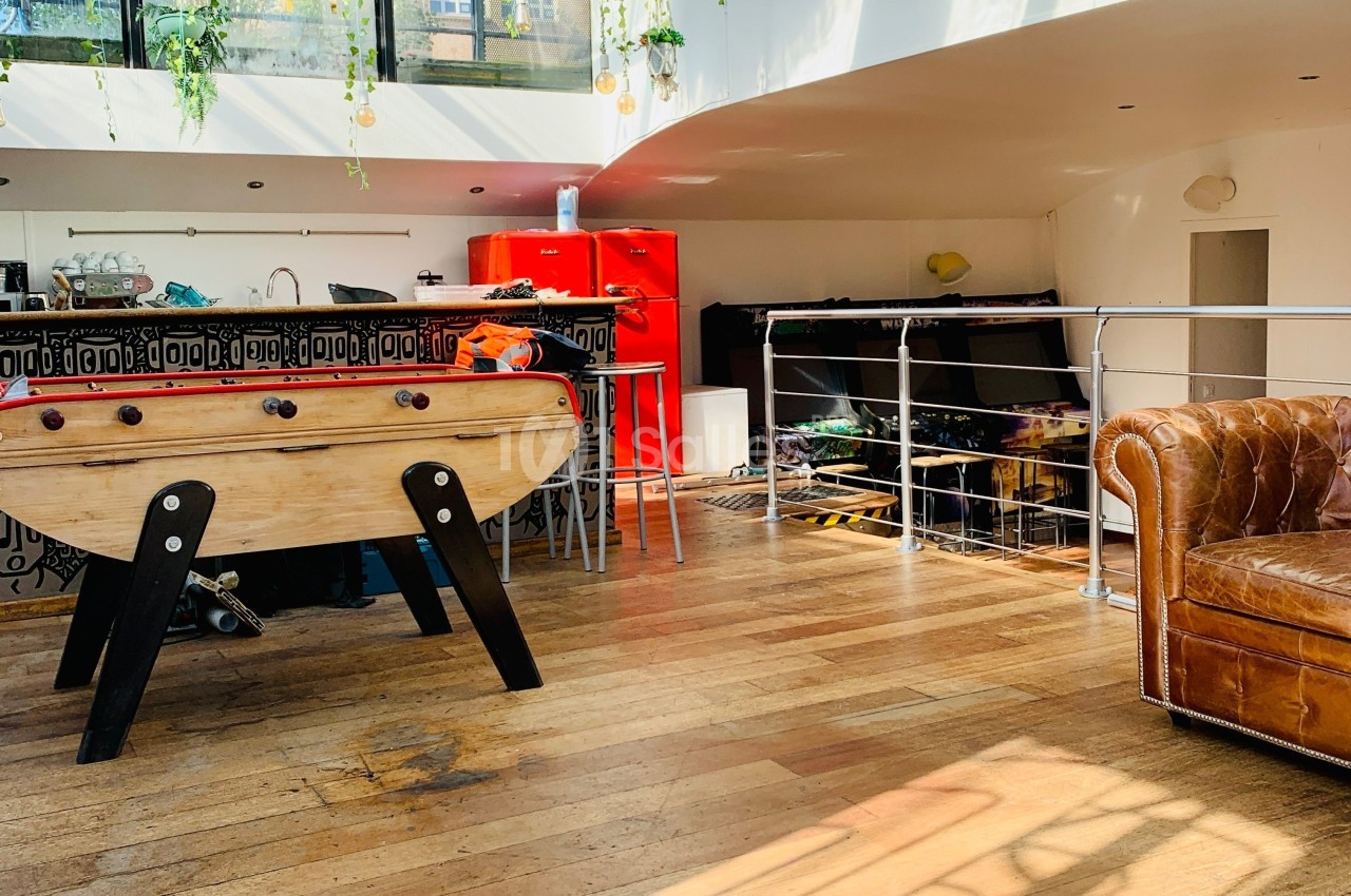 Espace lumineux avec baby-foot, canapé en cuir, cuisine ouverte et mezzanine, parquet en bois au sol.